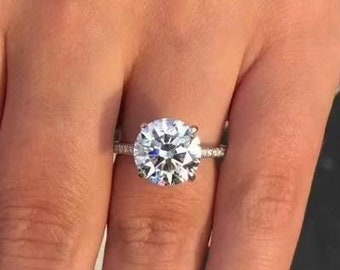 Könnte beinhalten: Nahaufnahme einer Hand, die einen silbernen Ring mit einem großen, runden, klaren Diamanten trägt. Der Ring hat ein dünnes Band mit kleinen Diamanten, die um den Hauptstein gesetzt sind.