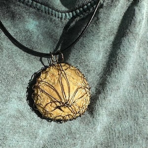 May include: A round pendant necklace with a beige stone center and a wire-wrapped design. The pendant hangs from a black cord. The person is wearing a blue-green t-shirt.