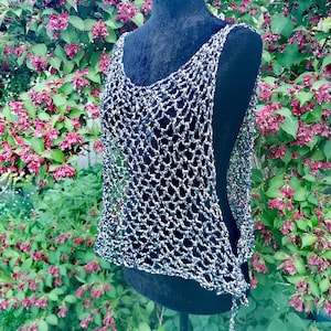 May include: A silver and black open-weave top displayed on a black mannequin. The top has a scoop neck and a loose, flowing design. The background features a vibrant bush with pink flowers and green foliage.
