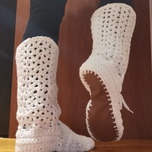 May include: White crocheted boots with a brown sole. The boots are made with a simple stitch pattern and are tall enough to reach the calf.