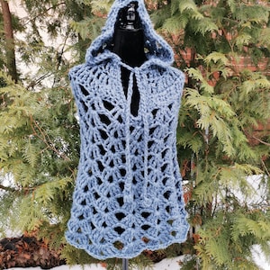 May include: A blue crocheted hooded vest with a delicate openwork pattern. The vest is displayed on a mannequin against a backdrop of evergreen trees and snow.