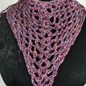 Puede incluir: Un chal de crochet hecho a mano, de tejido abierto, en tonos morados con acentos iridiscentes. La bufanda triangular se muestra en un maniquí negro, mostrando su delicado diseño calado. El hilo parece tener un brillo sutil.