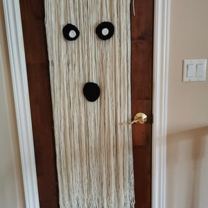 May include: A white yarn ghost wall hanging with black felt eyes and mouth. The ghost is hanging on a wooden door.