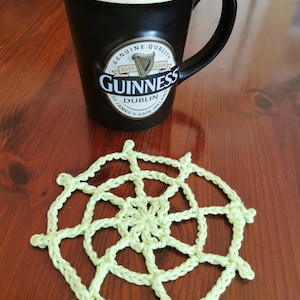 May include: A black Guinness mug with coffee on a wooden surface, next to a light green crocheted coaster. The mug has the Guinness logo and a white rim. The coaster has a circular, web-like design.