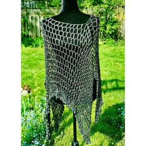 May include: A dark gray, open-weave crochet poncho featuring an asymmetrical hemline and a loose, net-like stitch pattern. The garment is displayed on a black mannequin, showcasing the intricate, textured yarn construction and the wide, draped neckline.