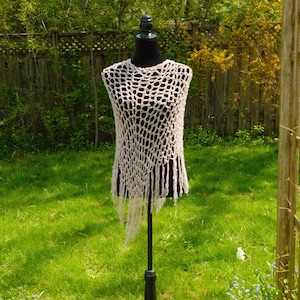May include: A cream-colored, open-weave crochet top with fringe, displayed on a black mannequin. The top is sleeveless and has a loose, airy design. The background is a green lawn and trees.