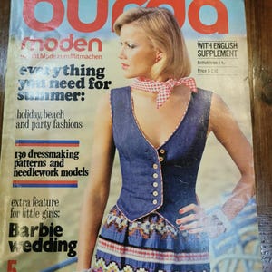 Peut inclure: Un magazine vintage Burda Moden de mai 1976 présentant en couverture une personne portant un gilet en jean bleu foncé avec des boutons blancs, un foulard à carreaux rouges et blancs et une jupe à motifs multicolores en zigzag.
