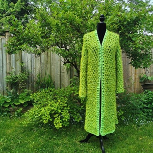 May include: A long, lime green crocheted cardigan with a front opening and a crocheted trim. The cardigan is displayed on a black mannequin against a green grassy background.