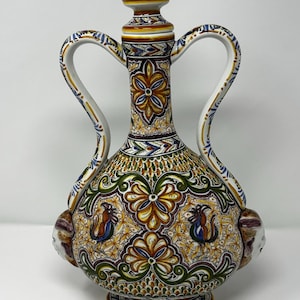 May include: A decorative ceramic pitcher with a bulbous base, two curved handles, and a conical stopper. The pitcher features intricate floral and geometric patterns in shades of brown, yellow, green, and blue. The handles are white with blue and yellow accents.