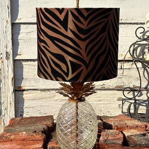 Könnte beinhalten: Dekorative Tischlampe mit einem Sockel aus klarem Glas in Ananasform und einem goldfarbenen Ananasaufsatz. Der Lampenschirm hat ein braunes und schwarzes Zebra-Streifenmuster. Die Lampe steht auf einem Stapel roter Ziegelsteine.