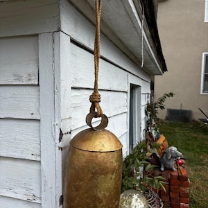 Puede incluir: Una gran campana de latón de estilo antiguo cuelga de un gancho en una estructura de madera blanca. La campana tiene una parte superior redondeada y un cuerpo cilíndrico, mostrando signos de desgaste. La cuerda es gruesa y anudada.