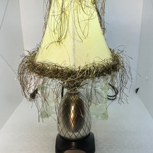 Puede incluir: Una lámpara de mesa con forma de piña con una pantalla de color crema ribeteada con flecos marrones.