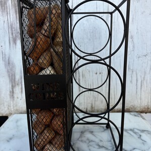 May include: Black metal wine rack with a section for wine corks and space for four bottles. The cork section is a mesh cage filled with used corks. The words "WINE CORKS" are cut out of the metal.