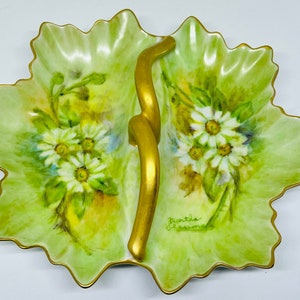 May include: A green and gold leaf-shaped serving dish with a hand-painted floral design of white daisies. The dish has a gold handle and is signed "Martha Jackson".
