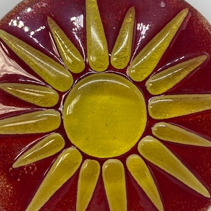 Puede incluir: Un atrapasol redondo de vidrio rojo con un diseño de sol amarillo. El sol tiene 12 rayos y un centro amarillo.