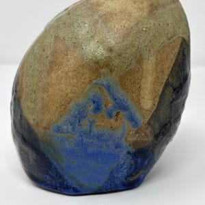 May include: A handcrafted ceramic vase with a unique, organic shape. The vase features a blend of colors, including brown, tan, blue, and black, creating a textured, earthy appearance. The vase has a small opening at the top.