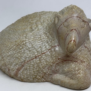 May include: A carved stone sculpture of a bear in a resting position. The bear is light gray with reddish-brown veining and a textured surface. The sculpture has a detailed face and ears.