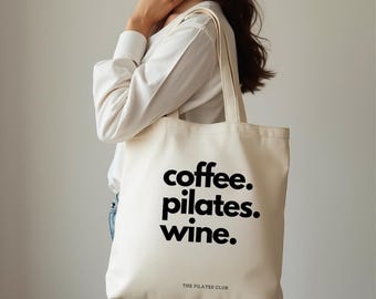 Puede incluir: Bolso tote de color crema con las palabras "coffee. pilates. wine." en negrita negra. El texto "THE PILATES CLUB" está impreso debajo. El bolso lo lleva una persona.