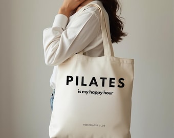 May include: Cream-colored canvas tote bag with the words "PILATES is my happy hour" in bold black letters. The Pilates Club is printed at the bottom. The bag has a simple design and is carried by a person.