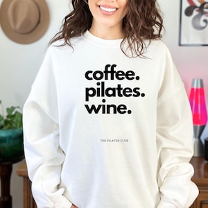 Peut inclure: Un sweat-shirt blanc avec les mots "coffee. pilates. wine." en gras et en noir. En dessous, le texte "THE PILATES CLUB" est écrit en plus petit. Le sweat-shirt est de style décontracté.