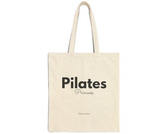 May include: Cream-colored tote bag with black text that reads "Pilates Princess" in a bold font, with "Princess" in a script font. The words "Pilates Club" are printed below. The bag has two long handles.