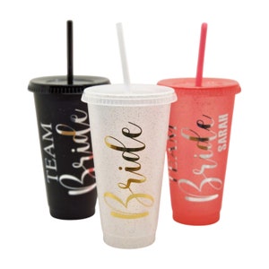 Könnte beinhalten: Drei wiederverwendbare Becher mit Deckel und Strohhalmen. Die Becher sind schwarz, weiß mit Glitzer und rosa. Jeder Becher hat das Wort "Bride" in goldenen Buchstaben darauf geschrieben. Der schwarze Becher hat auch die Worte "Team Bride" darauf geschrieben. Der rosa Becher hat den Namen "Sarah" darauf geschrieben.
