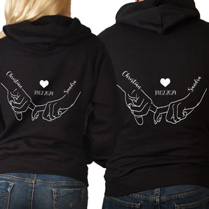 Könnte beinhalten: Zwei schwarze Hoodies mit weißem Text und einem Herz-Design. Der Text auf den Hoodies lautet "Christian" und "Sandra" mit dem Datum "14.02.2024". Das Design zeigt zwei Hände, die sich am kleinen Finger festhalten.