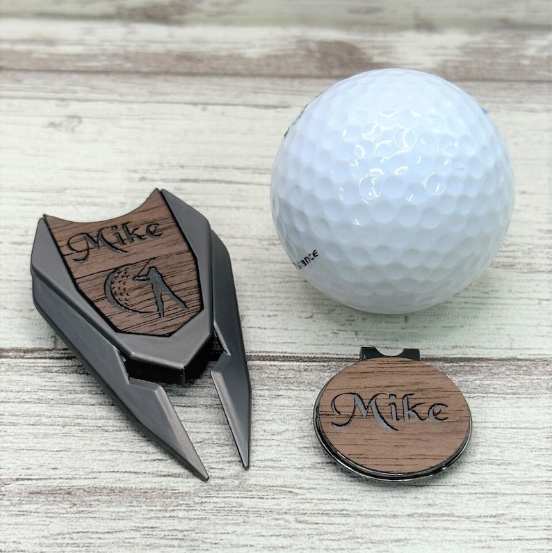 May include: Personalised golf ball marker and divot tool set with a wood grain finish. The set includes a black metal divot tool with a wood inlay featuring a golfer silhouette and the name "Mike" and a round wood ball marker with the name "Mike" engraved on it.