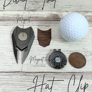 Pu&ograve; includere: Uno strumento per riparare i divot da golf con un fermaglio per cappello magnetico. Lo strumento per riparare i divot &egrave; in metallo e ha un accento in legno. Il fermaglio per cappello &egrave; anch'esso in metallo e ha un accento in legno. Il testo "Divot Tool" e "Hat Clip" &egrave; scritto in corsivo nell'immagine.
