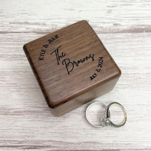 May include: A wooden ring box with the text "Kyle & Julie The Browns July 8, 2024" engraved on the top. Two wedding bands are sitting next to the box.