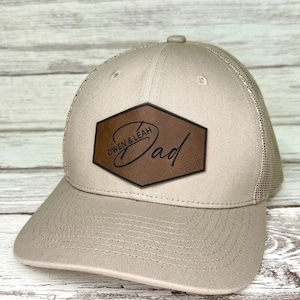 Peut inclure: Casquette de baseball beige avec un dos en filet. Un écusson en cuir marron sur le devant affiche le mot "Dad" en écriture cursive, avec "Owen & Leah" au-dessus. La casquette a une visière incurvée et un bouton sur le dessus.