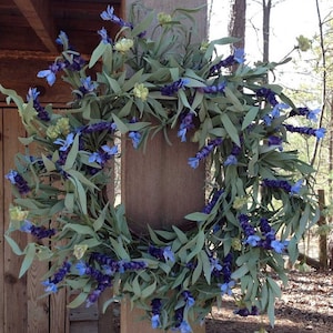 May include: A lavender wreath with green leaves and purple flowers. The wreath is hanging on a wooden post.
