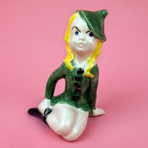 May include: A ceramic figurine of a girl wearing a green hat and dress. She has yellow hair and a serious expression.