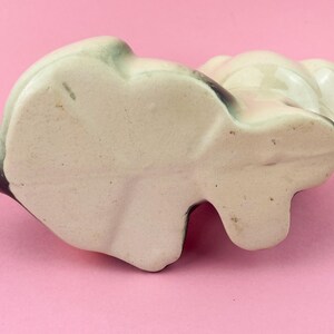 May include: A ceramic panda figurine, viewed from below, with a cream-colored body and black and green accents. The panda has a rounded shape with visible ears and a small, flat base. The figurine is on a pink surface.