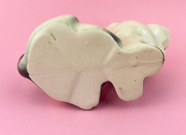 May include: A ceramic panda figurine, viewed from below, with a cream-colored body and black and green accents. The panda has a rounded shape with visible ears and a small, flat base. The figurine is on a pink surface.