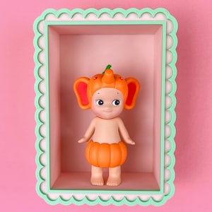 May include: A small, orange pumpkin-themed figurine with a smiling face, standing inside a pink rectangular box with a scalloped green frame. The figurine has a pumpkin head and skirt, with orange ears and a green stem. The background is a solid pink.