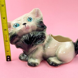 May include: A vintage ceramic cat planter, featuring a cream-colored body with black and gray accents. The cat has blue eyes and a pink nose. The planter measures approximately 5 inches in length.