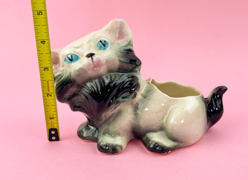 May include: A vintage ceramic cat planter, featuring a cream-colored body with black and gray accents. The cat has blue eyes and a pink nose. The planter measures approximately 5 inches in length.