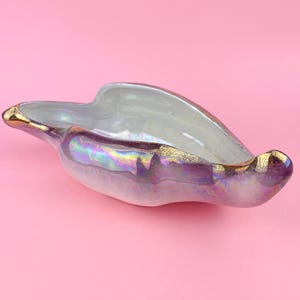 May include: An iridescent ceramic bowl with a unique, asymmetrical shape. The bowl features a pearlescent finish with hints of purple and gold accents along the rim. The interior is a light, neutral color.