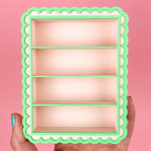 May include: A small, rectangular shelf with four levels. The shelf has a light pink interior and is framed with a scalloped, mint green border. The shelf is held against a pink background.