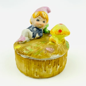 May include: A ceramic trinket box with a whimsical design featuring a girl with blonde hair wearing a pink hat and a blue dress, sitting next to a yellow mushroom with red spots. The box is decorated with a brown and green woodland scene.