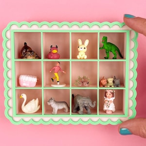May include: A miniature display case with a mint green scalloped frame, holding various small figurines. The collection includes a turtle, duck, bunny, dinosaur, human figure, teeth, swan, dog, elephant, and a doll. The background is pink.