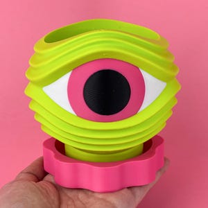 May include: A vibrant, eye-shaped decorative object in neon green, pink, white, and black. The object has a layered design with a large eye in the center. The base is a wavy pink shape. The background is a solid pink.