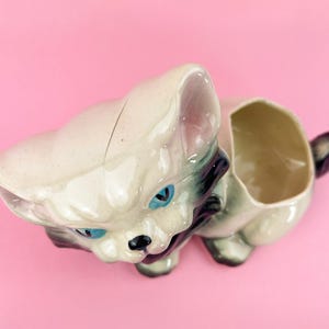 May include: A vintage ceramic cat planter in a cream color with blue eyes and black accents. The cat-shaped planter has an open compartment for holding small plants or other items. The tail is black and the ears are pink.