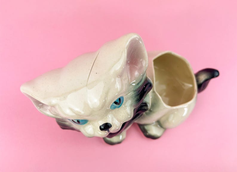 May include: A vintage ceramic cat planter in a cream color with blue eyes and black accents. The cat-shaped planter has an open compartment for holding small plants or other items. The tail is black and the ears are pink.