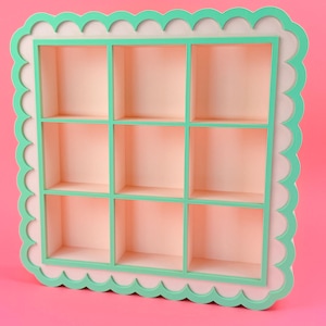 May include: A square wall shelf featuring nine equal compartments. The frame is light pink with a scalloped edge outlined in bright mint green. The interior dividers are also finished in mint green, creating a grid pattern against the light pink back.