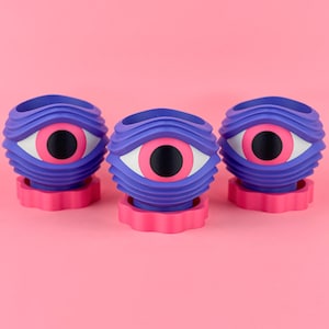 May include: Three eye-shaped decorative objects. Each has a purple, ridged body, a pink iris, white eyelids, and a black pupil. They sit on pink, scalloped bases against a pink background. Likely planters or small containers.