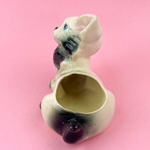 May include: A vintage ceramic dog planter in a sitting position, with a large opening for plants. The dog is primarily white with black and gray accents, including its ears and tail. The eyes are blue.