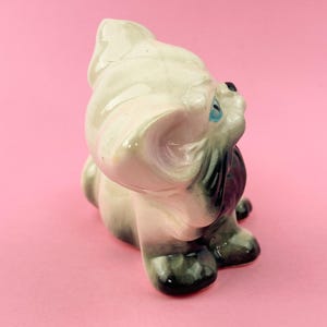 May include: A vintage ceramic figurine of a seated dog, primarily off-white with gray and black accents. The dog has blue eyes and a glossy finish. The figurine is set against a pink background.