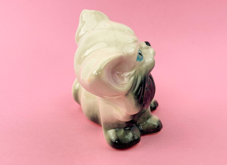 May include: A vintage ceramic figurine of a seated dog, primarily off-white with gray and black accents. The dog has blue eyes and a glossy finish. The figurine is set against a pink background.